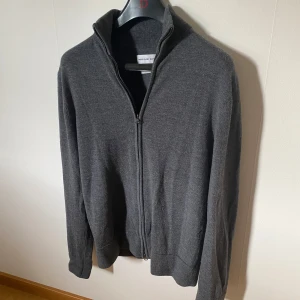 Mörkgrå merinoull kofta These Glory Days - Snygg mörkgrå kofta från These Glory Days i mjuk superior merinoull. Modellen har hel dragkedja, ribbad krage och muddar samt långärmad passform. Perfekt att slänga på över en t-shirt för en chill och stilren look.