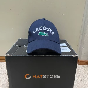 Mörkblå Lacoste keps - Snygg mörkblå keps från Lacoste med klassisk böjd skärm och broderad logga framtill. Kepsen har justerbar rem baktill och är tillverkad i bomull för en bekväm passform. Perfekt för att lyfta din streetwear-look.