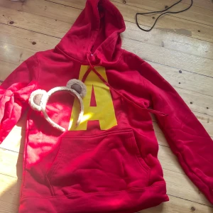 Röd hoodie med gul bokstav A - Säljer en röd hoodie med stor gul bokstav A på bröstet. Tröjan har huva med snörning och en stor magficka framtill. Perfekt för dig som vill klä ut sig till Alvin i gänget till halloween (öronen följer me)