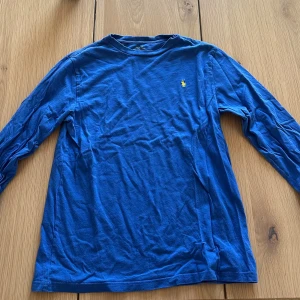 Blå långärmad tröja från Polo Ralph Lauren - Snygg blå långärmad tröja från Polo Ralph Lauren med klassisk gul logga broderad på bröstet. Tröjan är i mjuk bomull och har en rund hals. Perfekt för dig som gillar stilrena och enkla plagg med en touch av ikonisk design.