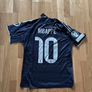 Real Madrid Mbappé matchtröja blå - Snygg Real Madrid fotbollströja med Mbappé och nummer 10 på ryggen. Tröjan är mörkblå med vita detaljer, Adidas-logga och klubbmärke på bröstet samt Champions League-märke på ärmen. Tillverkad i lätt och ventilerande material, perfekt för träning eller match.