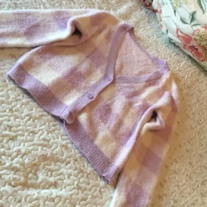 Ljuslila rutig kofta med knappar - Supermysig och fluffig kofta i ljuslila och beige rutor. Koftan har v-ringad hals, långa ärmar och stängs med tre matchande knappar framtill. Materialet är stickat och känns mjukt mot huden, perfekt för chilliga dagar. Croppad passform som ger en trendig vibe.