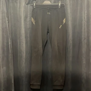 Svarta mjukisbyxor från Hugo Boss - Svarta mjukisbyxor från Hugo Boss med gulddetaljer vid fickorna och logga på ena benet. Byxorna har dragsko i midjan och är tillverkade i en mjuk bomulls- och polyestermix. Perfekta för en chill och snygg look.