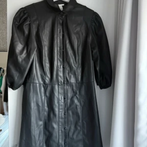 Svart skinnklänning H&M puffärm -  svart klänning i skinnimitation från H&M med puffiga halvlånga ärmar och knappar framtill. Rak passform och markerad midja. Få tal gånger använd inget anmärkningsvärt 