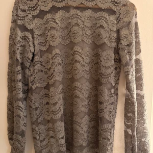 Grå/brun spetsblus från Soyaconcept - Supersnygg grå/brun långärmad blus i spets från Soyaconcept. Blusen har ett vackert blommigt spetsmönster, transparent tyg och en liten knapp i nacken. Perfekt för dig som vill sticka ut med en elegant och trendig look.