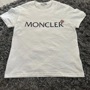 Vit Moncler t-shirt med logga - Vit t-shirt från Moncler med stor svart logotyp tryckt över bröstet och ett broderat Moncler-märke i vitt, blått och rött. Klassisk rund hals och korta ärmar. Tillverkad i mjuk bomull för skön känsla och clean look.