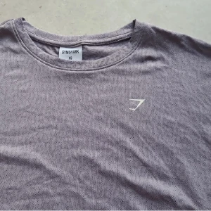 Lila croppad top från Gymshark - Säljer en ljuslila croppad t-shirt från Gymshark med rund halsringning och korta ärmar. T-shirten har en diskret logga på bröstet och är tillverkad i ett mjukt, stretchigt material. Perfekt för en sportig och avslappnad stil.