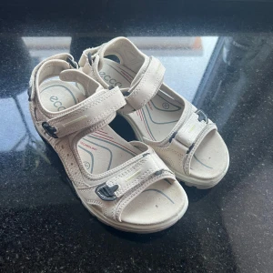 Beige sandaler från ECCO strl 36 - Snygga och bekväma sandaler från ECCO i beige skinn med justerbara kardborreband och svarta spänndetaljer. Sulan har bra grepp och är designad för komfort, perfekt för långa dagar på språng. Sportig look med öppen tå och diskreta kontrastdetaljer.
