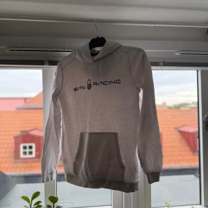 Vit hoodie från Sail Racing med tryck - Vit hoodie från Sail Racing med svart logotyptryck på bröstet. Tröjan har huva, stor magficka och ribbade muddar vid ärmslut och nederkant. Mjuk insida i bomullsmix, perfekt för chill eller häng. Snygg och enkel design som funkar till jeans eller joggers.