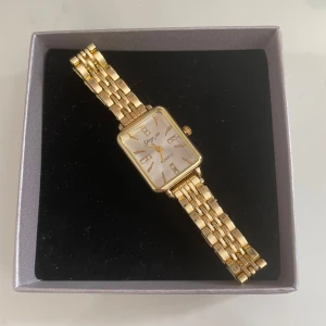 Guldig rektangulär klocka - Stilren klocka med rektangulärt ur och länkarmband i guld. Urtavlan är vit med gulddetaljer och små glittriga stenar vid vissa siffror. Perfekt accessoar för att lyfta din outfit. Vet ej vart den är ifrån därav priset.