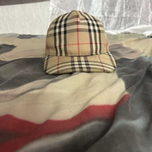 Burberry rutig keps med klassiskt mönster - Snygg keps från Burberry med det ikoniska rutiga mönstret i beige, svart, vitt och rött. Justerbar baktill med svart spänne och tydlig Burberry London England-patch. Perfekt accessoar för att lyfta din outfit med lyxig känsla.