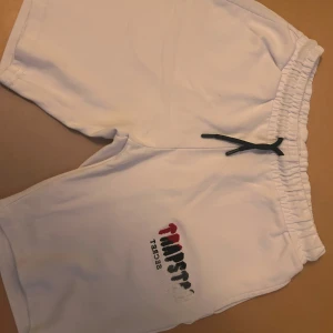 Vita Trapstar shorts med snörning - Snygga vita shorts från Trapstar med broderad logga i rött, svart och vitt på ena benet. De har elastisk midja med svart snörning för skön passform. Materialet är mjukt och känns som bomull, perfekt för chill dagar. Stilen är loose och midwaist. Storlek L men passar även S/M