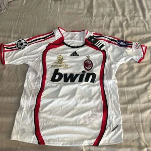 AC Milan Kaka 22 vit matchtröja Adidas - AC Milan fotbollströja med Kaka och nummer 22 på ryggen. Vit med röda och svarta detaljer, Adidas-logga och Champions League patch på ärmen. ClimaCool-material som andas, kortärmad och med snygga ränder längs axlarna. Perfekt för dig som älskar fotboll och retrostil.
