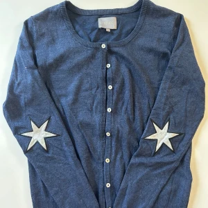Blå kofta med stjärnor från Culture - Mysig blå kofta från Culture med vita knappar framtill och coola silverfärgade stjärnor på ärmarna. Rund halsringning och ribbade muddar ger en snygg och enkel look. Perfekt för dig som vill sticka ut lite extra.