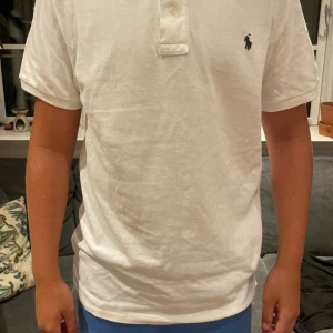 Vit pikétröja från Polo Ralph Lauren XL - Vit pikétröja från Polo Ralph Lauren i storlek medium/xl. Klassisk design med krage, korta ärmar och två knappar framtill. Ikoniska mörkblå Polo-loggan broderad på bröstet. Tillverkad i mjuk bomull som andas och känns skön mot huden.