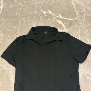 Svart pikétröja från Manfinity M - Svart pikétröja från Manfinity i storlek M. Tröjan har klassisk krage och korta ärmar, perfekt för en clean och stilren look. Materialet känns mjukt och lätt, vilket gör den skön att bära under varmare dagar.