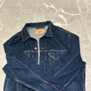 Levi's klassisk jeansjacka mörkblå L - Säljer en klassisk mörkblå jeansjacka från Levi's i modell 70550. Jackan har snygga kontrastsömmar, bröstfickor med knappar och den ikoniska röda Levi's-taggen. Perfekt för dig som gillar tidlös denimstil. KOM MED BUD