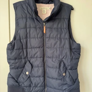 Mörkblå dunväst från H&M - Snygg mörkblå dunväst från H&M med quiltad design och hög krage. Västen har två fickor framtill med lock och tryckknapp samt dragkedja i mitten. Perfekt att slänga över en hoodie för extra värme under kyliga dagar.