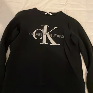 Svart Calvin Klein Jeans sweatshirt - Säljer en svart sweatshirt från Calvin Klein Jeans med stor vit CK-logga och text på bröstet. Tröjan har rund hals och långa ärmar, perfekt för en chill och stilren look. Materialet är mjukt och skönt, passar dig som gillar klassiska märken.