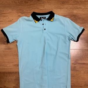 Fendi polo baby blue - Snygg ljusblå polo t-shirt från Zegna med svart krage och detaljer i gult. T-shirten har korta ärmar med svarta muddar och gula inslag, samt tre knappar framtill. Perfekt för dig som gillar stilrena och sportiga plagg.