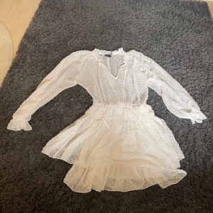 Vit kortklänning från ASOS DESIGN - Supersöt vit kortklänning från ASOS DESIGN med volangkant, långärmad och v-ringad. Klänningen har ett prickigt mönster i tyget, smockad midja och transparenta ärmar. Perfekt för dig som gillar romantisk och luftig stil.