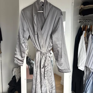 Grå omlott morgonrock Homewear - Mjuk och lyxig grå morgonrock från Homewear Collection. Omlottmodell med bälte i midjan och bred krage. Tillverkad i glansigt och lent material som känns riktigt skönt mot huden. Perfekt för chill dagar hemma.