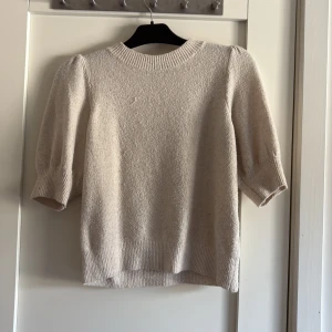 Beige stickad tröja Vero Moda XS - Supersöt beige stickad tröja från Vero Moda i storlek XS. Tröjan har puffiga korta ärmar, ribbade muddar och rund halsringning. Perfekt för dig som gillar en enkel men trendig look med lite volym på axlarna.