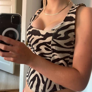 Zebramönstrad korsett från H&M Divided - Snygg zebramönstrad korsett-topp från H&M Divided i svart och beige med glittriga inslag. Modellen är croppad med breda axelband och markerade sömmar för en cool siluett. Perfekt för dig som vill sticka ut med ett djurmönster!