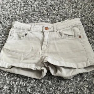 Ljusbeige jeansshorts med uppvik - Snygga ljusbeige jeansshorts med klassisk femficksdesign och uppvikta ben. De har knapp och dragkedja framtill samt bälteshällor. Perfekta för varma dagar och har en clean look som är lätt att matcha med olika stilar.