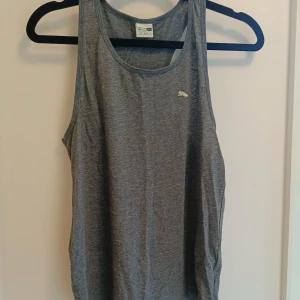 Grå linnetopp från Puma - Sportig grå linnetopp från Puma med klassisk logga på bröstet. Perfekt för träning eller chill dagar, tillverkad i mjukt och lätt material som andas. Ärmlös design med rund halsringning för maximal rörelsefrihet.