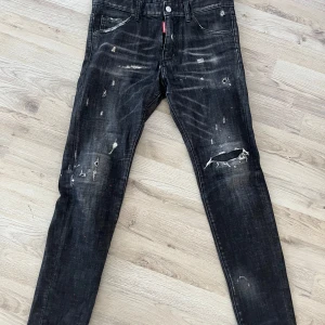 Svarta  jeans  Dsquared2 - Säljer ett par svarta jeans från Dsquared2 med coolt distressed och slitna detaljer, inklusive hål och färgstänk. Modellen har en smal passform och klassisk femficksdesign. Jeansen är tillverkade i jeansmaterial och har en röd Dsquared2-tag vid gylfen. Äkta 