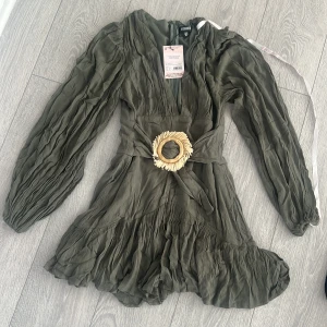 Olivgrön klänning med volang och bälte - Snygg olivgrön klänning från Mossman med volangdetaljer och ett dekorativt bälte med rund spänne. Klänningen har långa ärmar och en v-ringad design som ger en elegant touch. Perfekt för att sticka ut med stil!