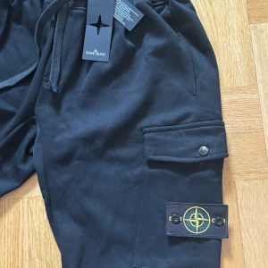 Svarta cargopants från Stone Island - Snygga svarta cargopants från Stone Island med praktiska fickor och justerbar midja med snörning. Byxorna har en ikonisk logga på benet och knappar med märkets namn. Perfekta för en stilren och avslappnad look.