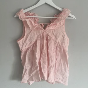 Rosa ärmlös blus med volangdetaljer - Söt rosa blus med volangdetaljer runt halsen och knytning framtill. Perfekt för en somrig look. Strl 170