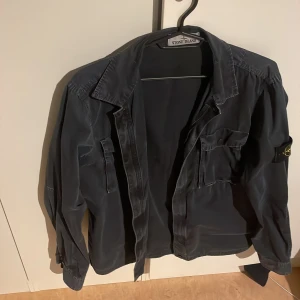 Svart overshirt från Stone Island - Säljer en stilren svart overshirt från Stone Island med deras ikoniska broderade märke på ärmen. Jackan har långa ärmar och en klassisk krage. Perfekt för en casual look. Äkta skick 9/10 Inga defekter bara lite urtvättad. Kom dm för mer frågor storlek L men passar också M