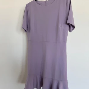 Ljuslila klänning med volangdetalj - Säljer en söt ljuslila klänning med korta ärmar och volangdetalj vid fållen. Klänningen har en rund hals och en dragkedja i ryggen för enkel påklädning. Perfekt för vårens och sommarens tillfällen!