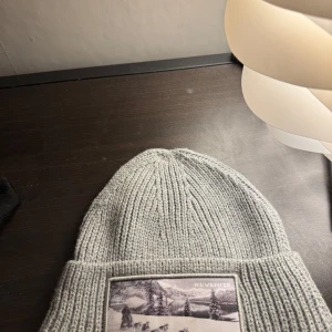 Grå stickad mössa med patch - Snygg grå stickad mössa med en cool patch framtill. Perfekt för att hålla värmen under kyliga dagar. Enkel och stilren design som passar till det mesta. Från revenite