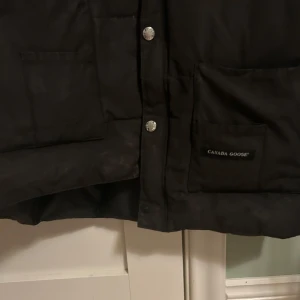 Svart dunväst från Canada Goose - Säljer en svart dunväst från Canada Goose med tryckknappar och två stora fickor framtill. Västen har en hög krage och det ikoniska Canada Goose-emblemet på bröstet. Perfekt för kyliga dagar!