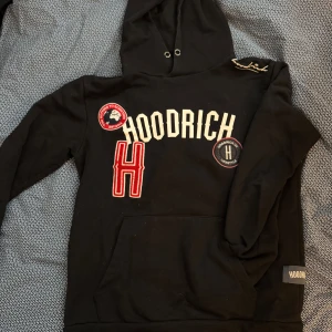 Svart hoodie från Hoodrich - Säljer en svart hoodie från Hoodrich med broderade detaljer och logotyp på bröstet. Tröjan har en stor ficka framtill och en justerbar huva. Perfekt för en avslappnad stil.