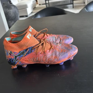 Puma future 8/elit - Kostar 2600 nypris, Säljer ett par unika fotbollsskor i en mix av orange, blå och lila med snörning. Skorna har en iögonfallande design med mönster och en hög ankelkrage för extra stöd. Perfekta för den som vill sticka ut på planen! 