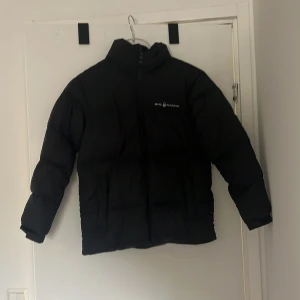 Svart dunjacka från Sail Racing - Säljer en svart dunjacka från Sail Racing. Jackan har en hög krage och dragkedja framtill. Perfekt för kalla dagar med sin pufferdesign som håller värmen. Märkeslogga synlig på bröstet och baksidan.