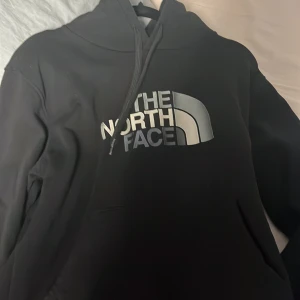 Svart hoodie från The North Face - Säljer en svart hoodie från The North Face med deras ikoniska logga i vitt och grått på bröstet. Tröjan har en klassisk design med huva och dragsko. Perfekt för en avslappnad stil.