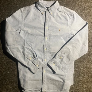 Ljusblå skjorta från Polo Ralph Lauren - Säljer en klassisk blå skjorta från Ralph Lauren med button-down krage och broderad logotyp på bröstet. Skjortan passar perfekt om du är cirka 155-165 cm lång. Om ni har några frågor är de bara att fråga på! Pris kan absolut diskuteras.