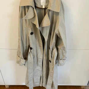 Beige trenchcoat från H&M - Snygg beige trenchcoat från H&M med klassisk design. Jackan har långa ärmar och knappar framtill samt ett bälte i midjan för en justerbar passform. Perfekt för vår och höst!
