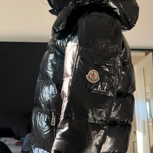 Maya Moncler Jacka - Snygg svart pufferjacka från Moncler med glansig yta och klassisk logga på ärmen. Jackan har en långärmad design med dragkedja. Den quiltade stilen ger extra värme och komfort. Säljer då den inte kommer till användning. Är i storlek 3 som motsvarar M. Se bild 2 för Certilogi-lappen.