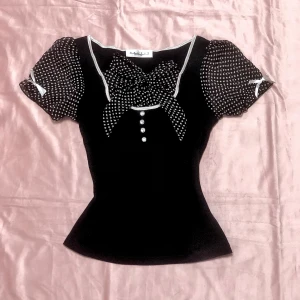 Svart prickig blus med rosett - Charmig svart blus med vita prickar och en stor rosett framtill. Blusen har korta puffärmar med små vita rosetter och dekorativa knappar. Perfekt för en söt och stilren look.                          Köpte för 75USD