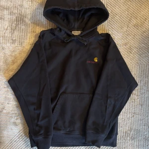 Svart hoodie från Carhartt - Säljer en svart hoodie från Carhartt med broderad logga i gult och rött på bröstet. Tröjan har en klassisk känguruficka och justerbar huva med snörning. Perfekt för en avslappnad stil. Har knappt användt den, och säljer pågrund av för liten storlek!