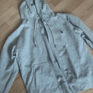 Grå hoodie från Ralph Lauren - Säljer en stilren grå hoodie från Ralph Lauren med dragkedja och justerbar huva. Den har en liten broderad logga på bröstet och praktiska fickor framtill. Perfekt för en avslappnad look. (Kan även byta) stl S men är som M 