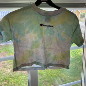 Tie-dye croppad t-shirt från Champion - Säljer en cool tie-dye croppad t-shirt från Champion. T-shirten har korta ärmar och ett broderat Champion-märke på bröstet. Perfekt för en avslappnad stil!