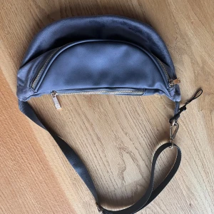 Grå magväska med dragkedja - Snygg blå magväska/axelväska med justerbar rem och dragkedja. Väskan har en stilren design med ett litet metallmärke på framsidan. Lappen är avtagen men den är helt oanvänd och i bra skicks.